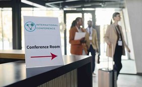 Zur gewerbesteuerlichen Hinzurechnung von Hotelzimmermieten bei einem Veranstalter für Konferenzen, Events und Reisen Ein Schild weist auf den Konferenzraum in einem Hotel. Im Hintergrund sind Teilnehmer zu sehen.